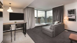 A modern hotel room with a dining area, a sofa, and a large window offering a city view. - Adina Apartment Hotel Hamburg Speicherstadt (Hamburg)