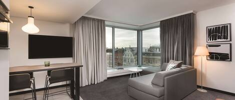 A modern hotel room with a dining area, a sofa, and a large window offering a city view.