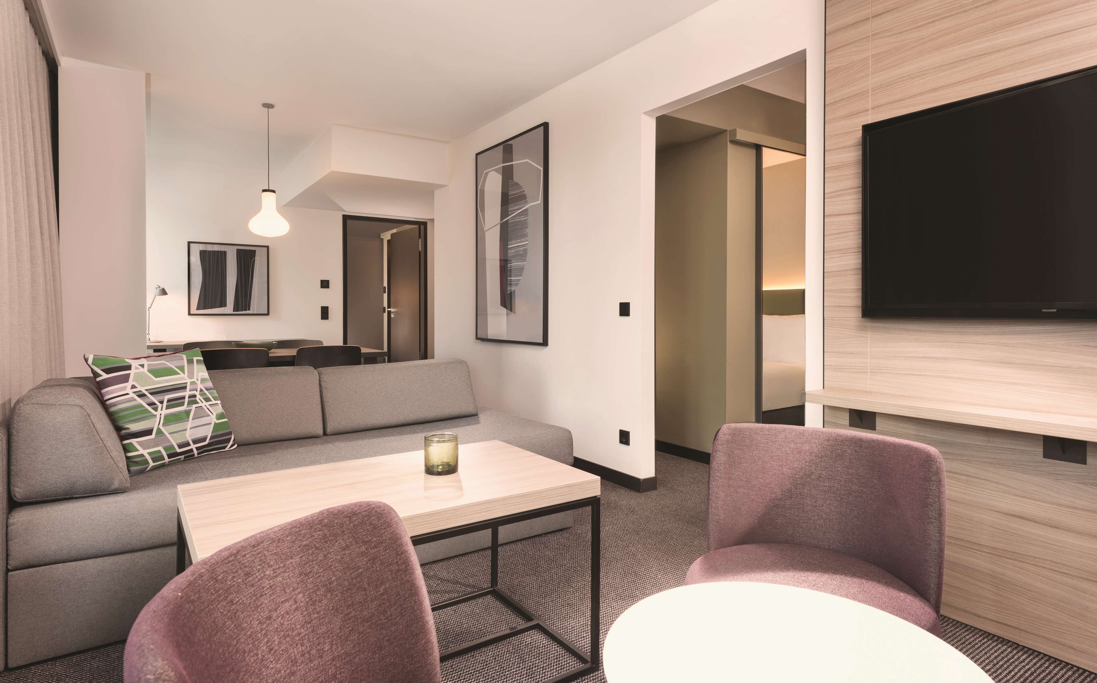 A modern hotel room with a sofa, two armchairs, a small table, a TV, and a painting.