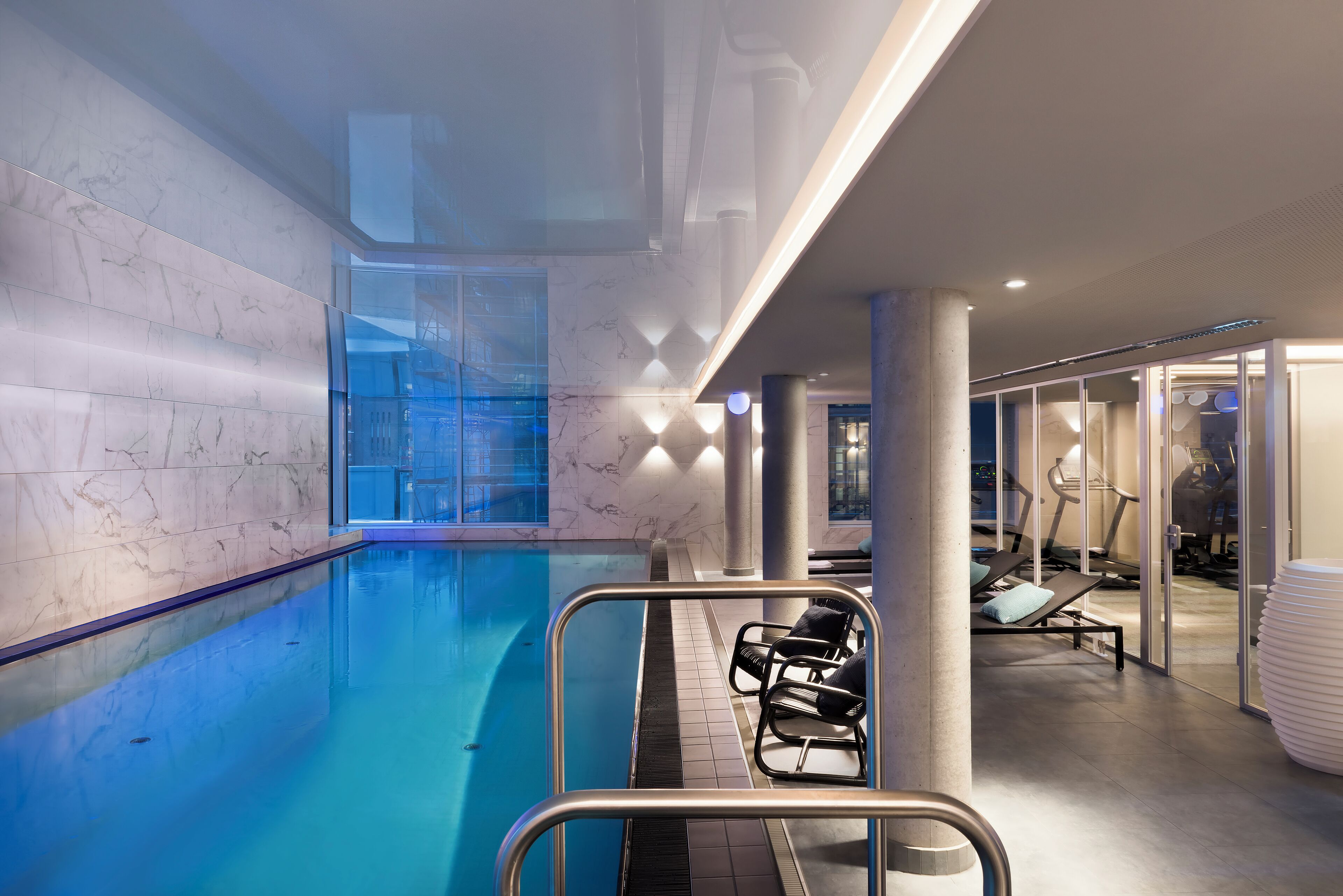 Una piscina interior con pared de mármol, cerramiento de vidrio y sillones de descanso.