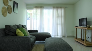 A living room with a grey sofa, a wooden TV stand, and a large window with sheer curtains. - Eagle Views Windhoek (Windhoek)
