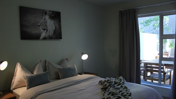 A bedroom with a bed, pillows, a blanket, a nightstand, a lamp, and a window with a view of outdoor furniture. - Eagle Views Windhoek (Windhoek)