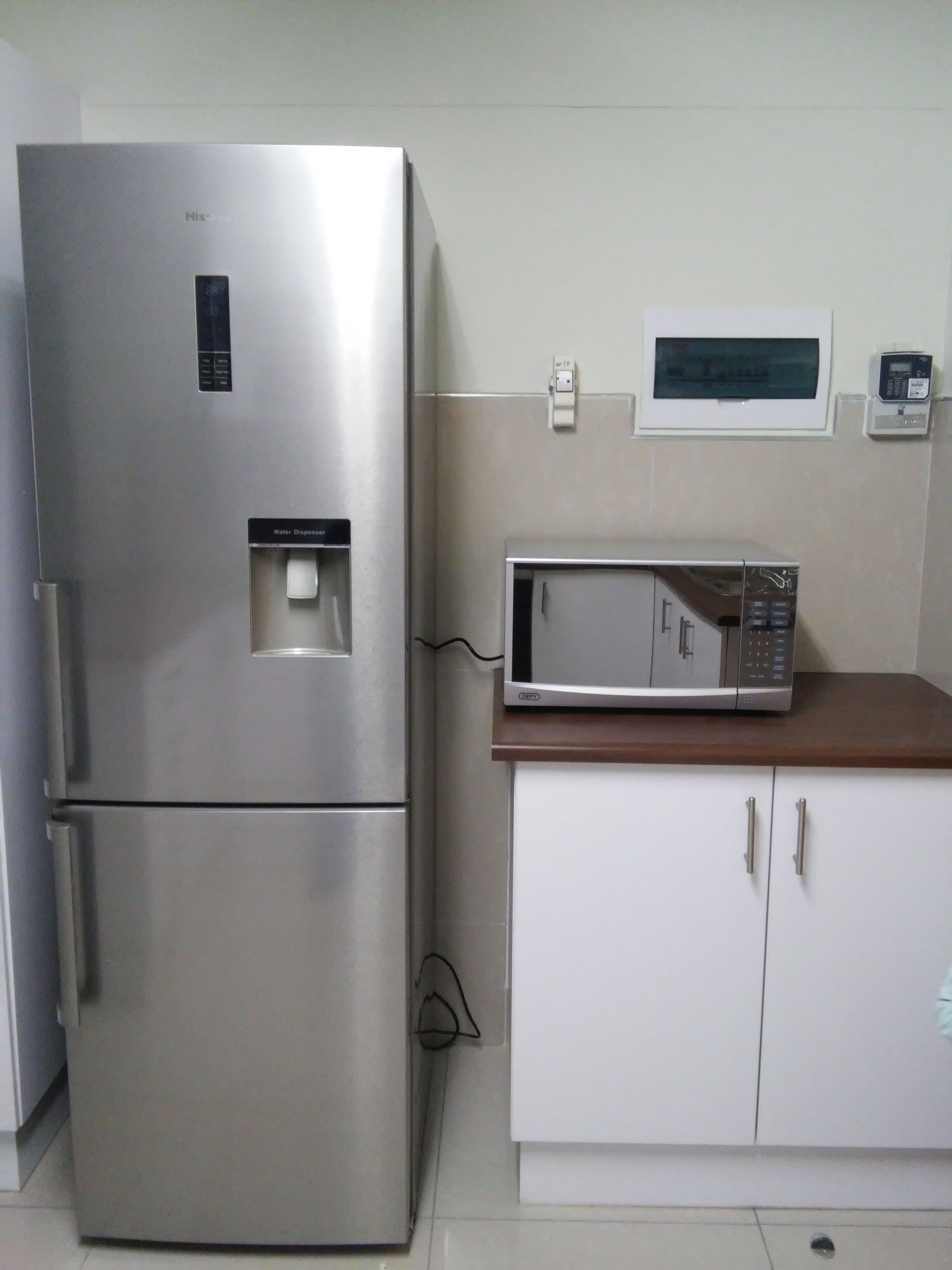 A stainless steel refrigerator with a water dispenser, a microwave on a wooden countertop, and a white cabinet with two drawers.