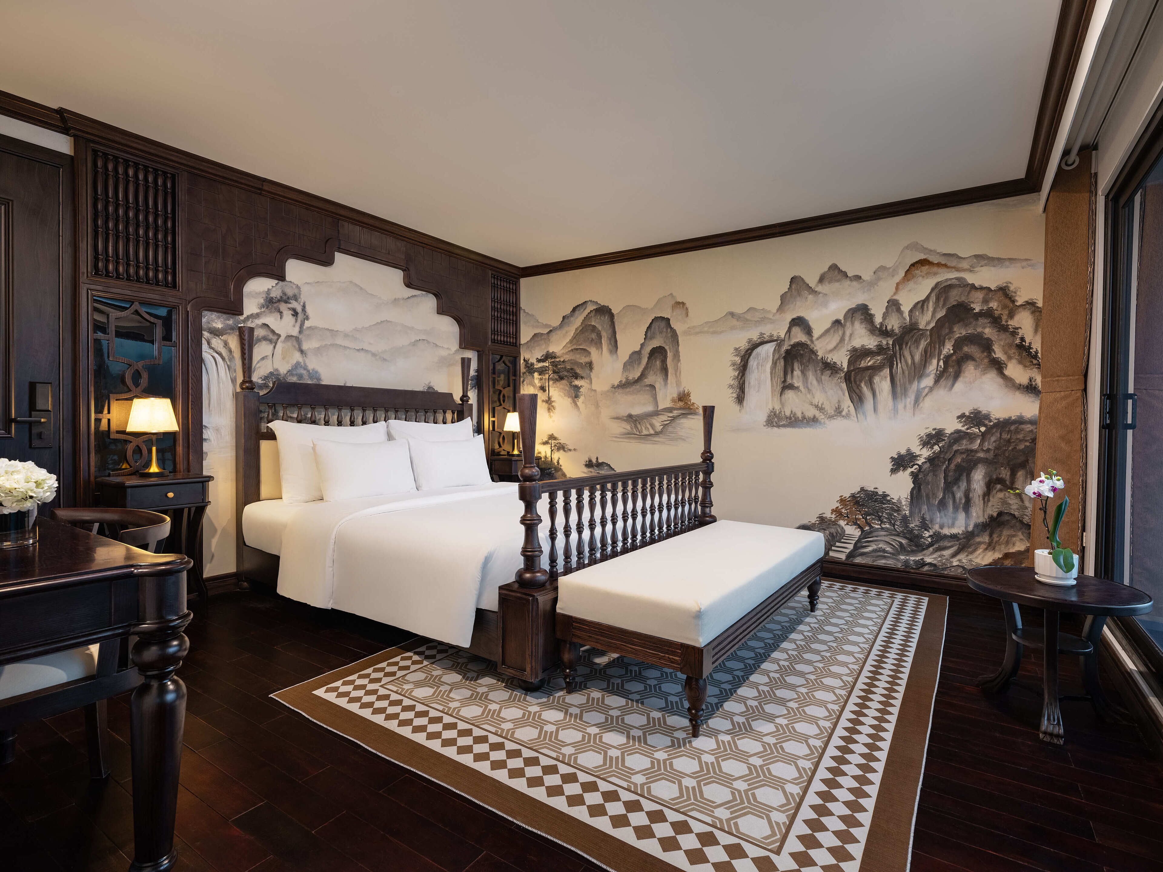 A traditional bedroom with a bed, a nightstand, a lamp, a wall mural, and a small table with a vase.