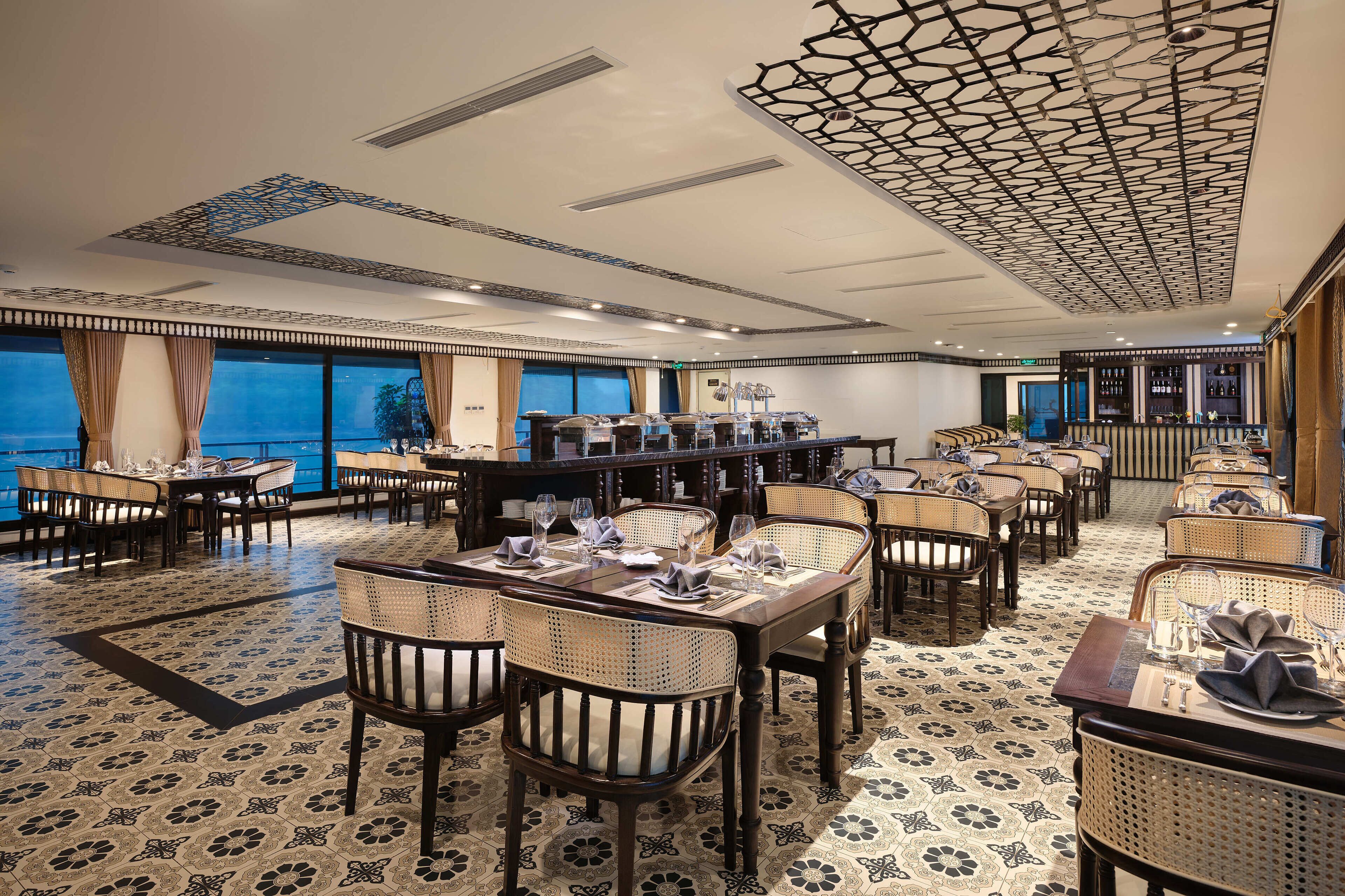 A spacious dining area with high tables and wicker chairs, large windows offering a view, and a patterned floor.