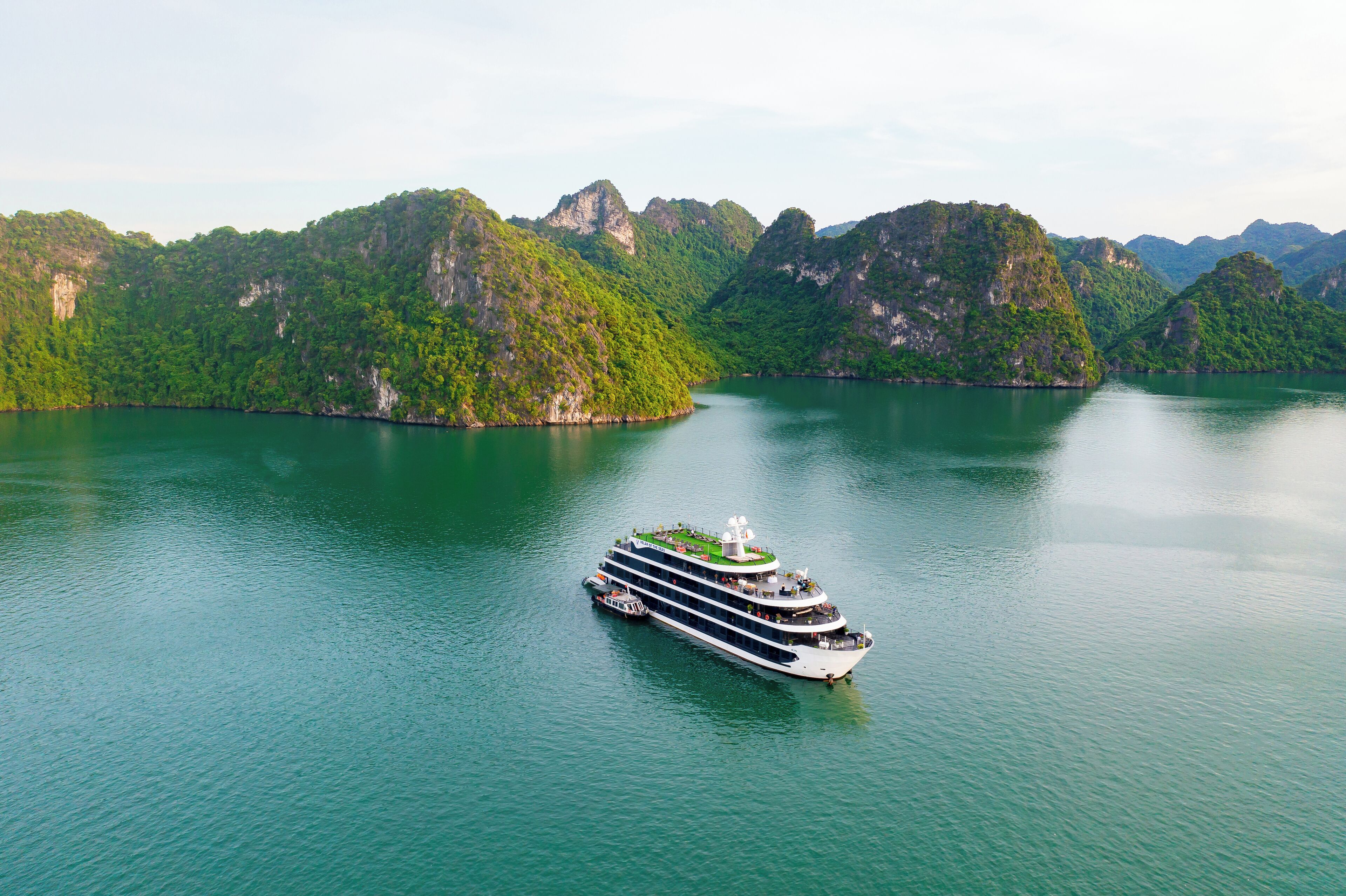 Un gran crucero con varias cubiertas y techo verde, navegando en un río tranquilo y verde, rodeado de colinas exuberantes y verdes.