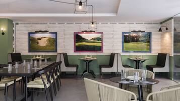 Un espace repas comprenant une longue table et des chaises, une petite table ronde, ainsi que trois tableaux représentant des paysages, encadrés et accrochés au mur.