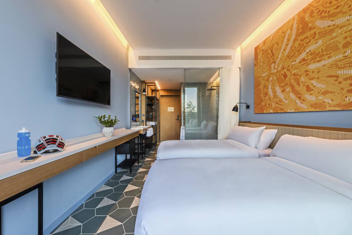 A modern hotel room with a large bed, a desk with a chair, a TV mounted on the wall, and a large abstract painting.