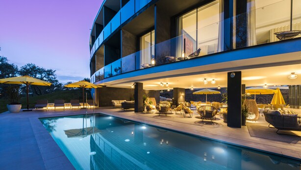 A modern outdoor pool area with lounge chairs and umbrellas, surrounded by a multi-story building with large windows and outdoor lighting.