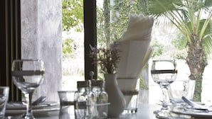 A dining table set with clear glassware and a white napkin folded on a plate, with a vase of flowers and a palm tree visible through the window. - Calvary Home (Mae Rim)
