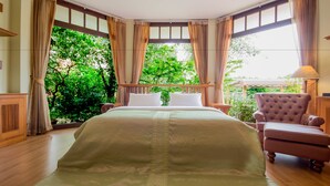 A bedroom with a large bed, a chair, and a view of greenery through the windows. - Calvary Home (Mae Rim)