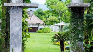 A garden entrance with a sign, lush greenery, and a thatched-roof structure in the background. - Calvary Home (Mae Rim)