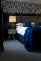 A hotel room with a bed, a nightstand with a lamp, patterned wallpaper, and a dark-colored headboard.