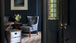 Una habitación azul oscuro con suelo de madera, un sillón estampado, una pequeña mesa con un florero y una maleta antigua.