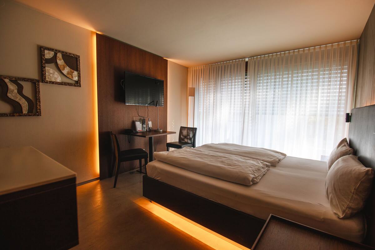 A modern hotel room with a large bed, a desk, a chair, and a TV mounted on the wall.