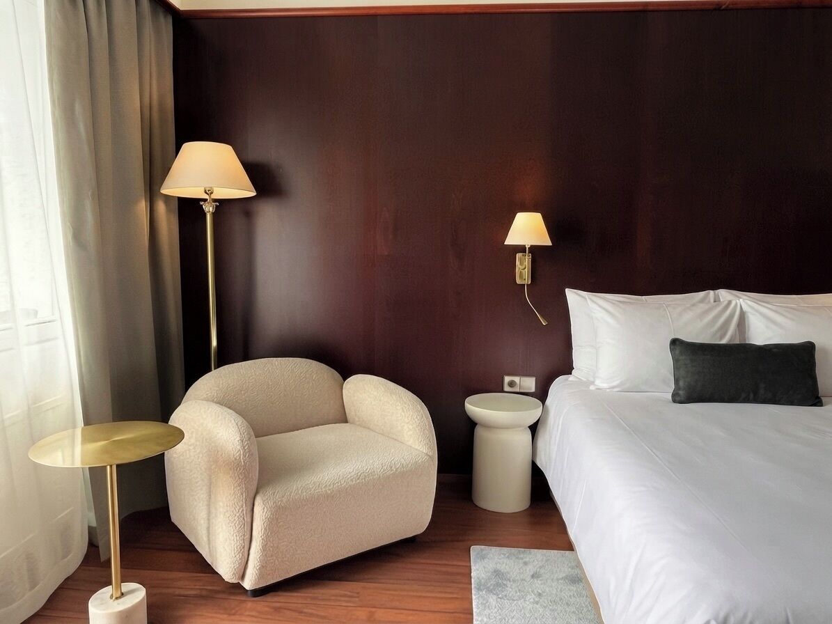 Una camera d'hotel con un letto, una sedia, un tavolino rotondo e due lampade a parete.