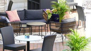 Een terras buiten met rieten meubels, waaronder een bank, stoelen en een tafel. Er staan potplanten en een bijzettafeltje met twee potplanten.