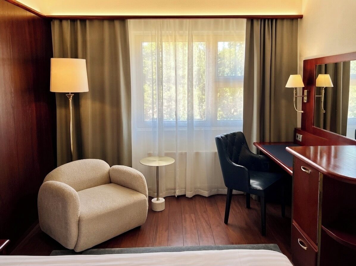 A hotel room with a bed, a desk, a chair, a lamp, a mirror, and a window with curtains.