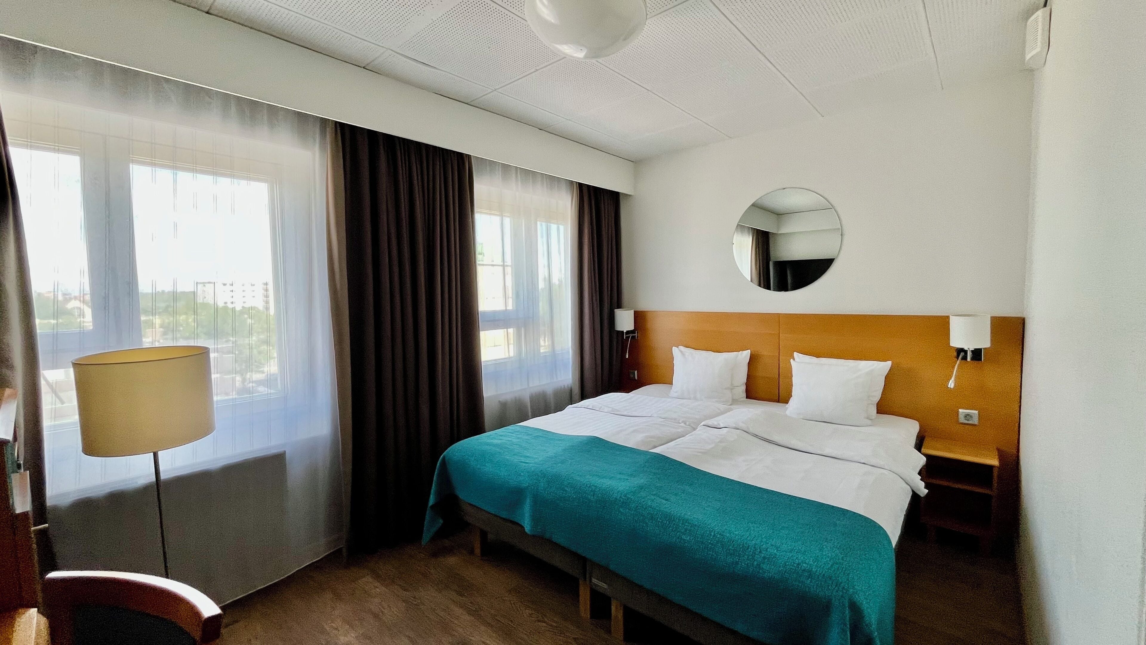 A hotel room with a bed, a bedside table, a lamp, a mirror, and a window with curtains.