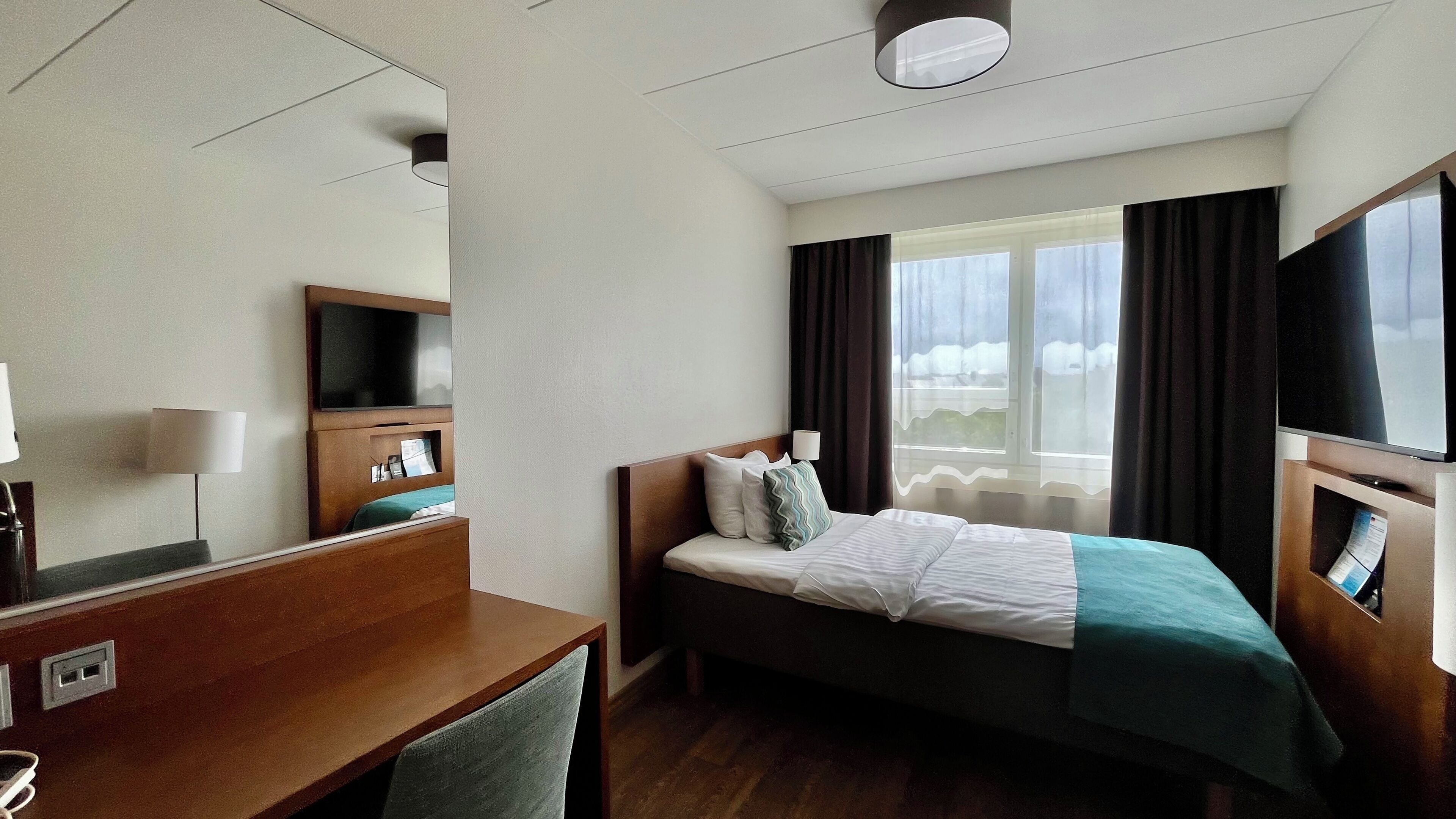 A hotel room with a bed, a desk, a TV, a window with curtains, and a ceiling light.