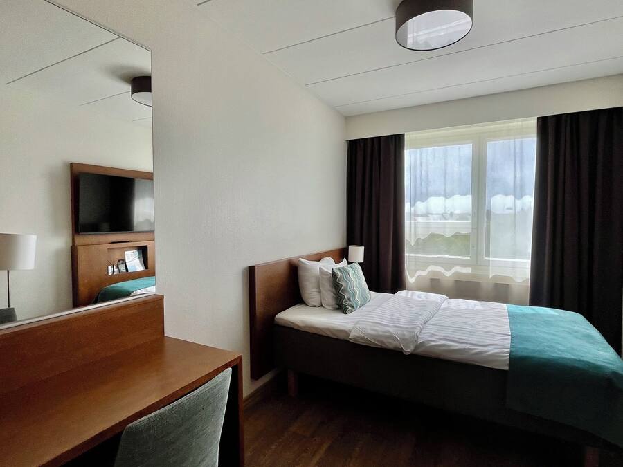 A hotel room with a bed, a desk, a TV, a window with curtains, and a ceiling light.