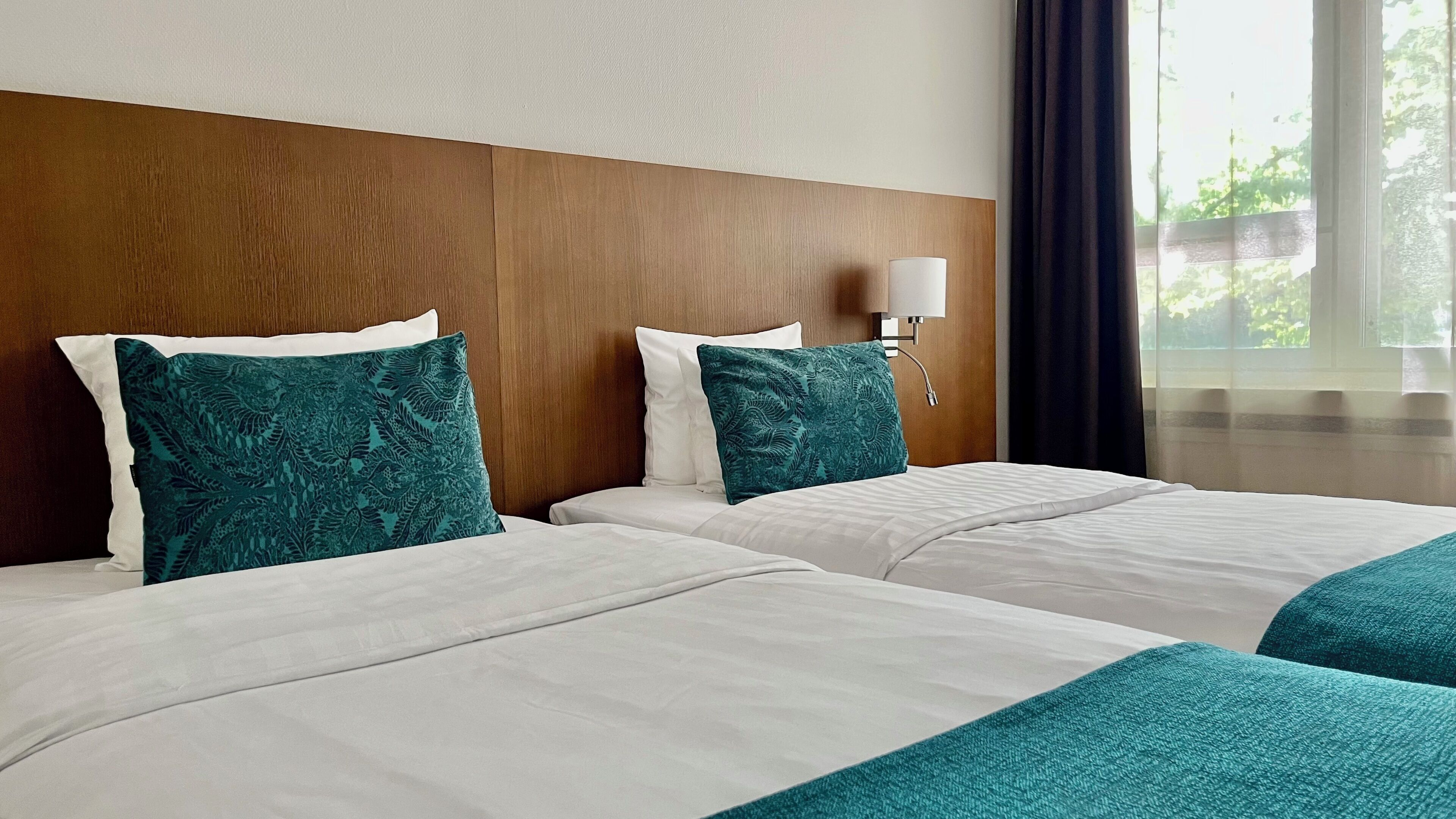 Habitación de hotel con dos camas, cabecero de madera, lámpara de noche y ventana con cortinas.