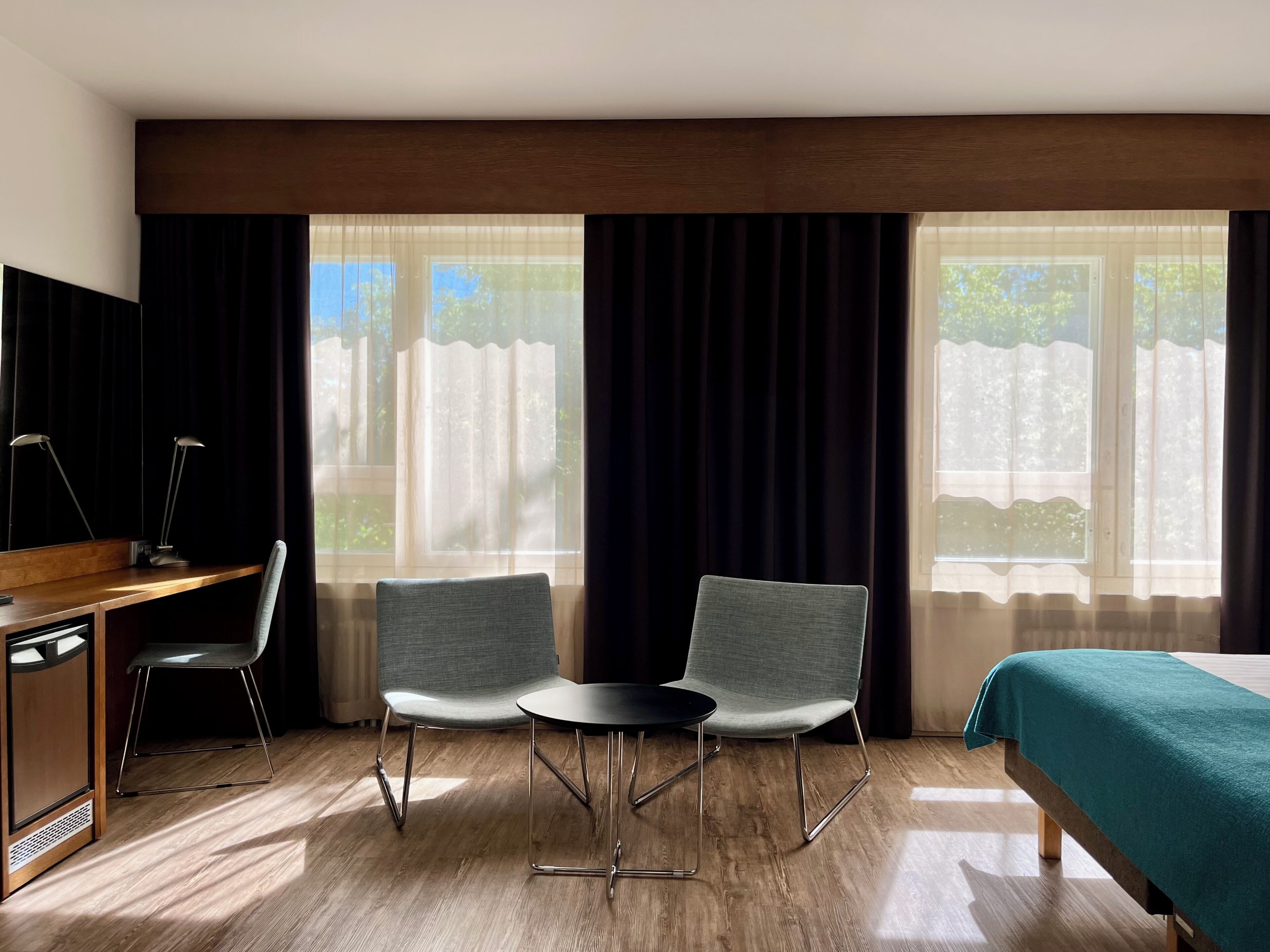 A hotel room with a wooden desk, two chairs, a small table, a bed with a green bedspread, and large windows with curtains.