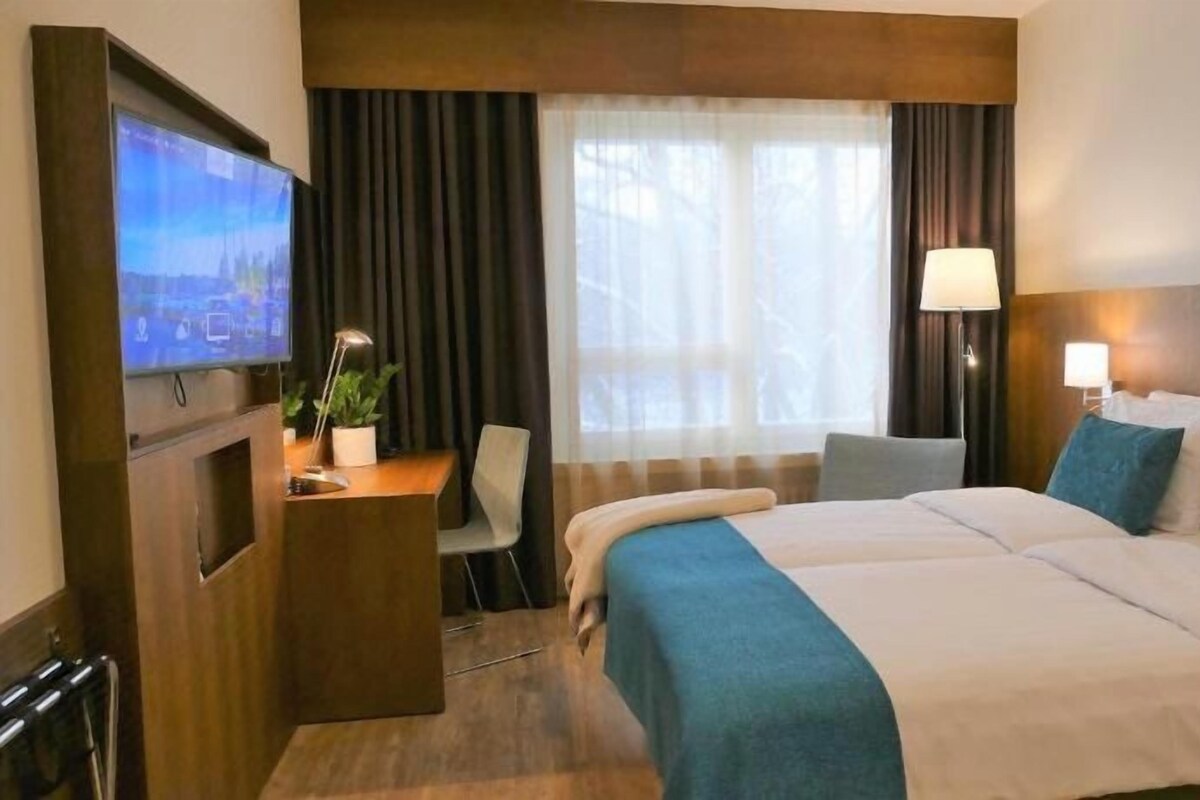 A hotel room with a bed, a desk with a computer, a TV, and a window with curtains.
