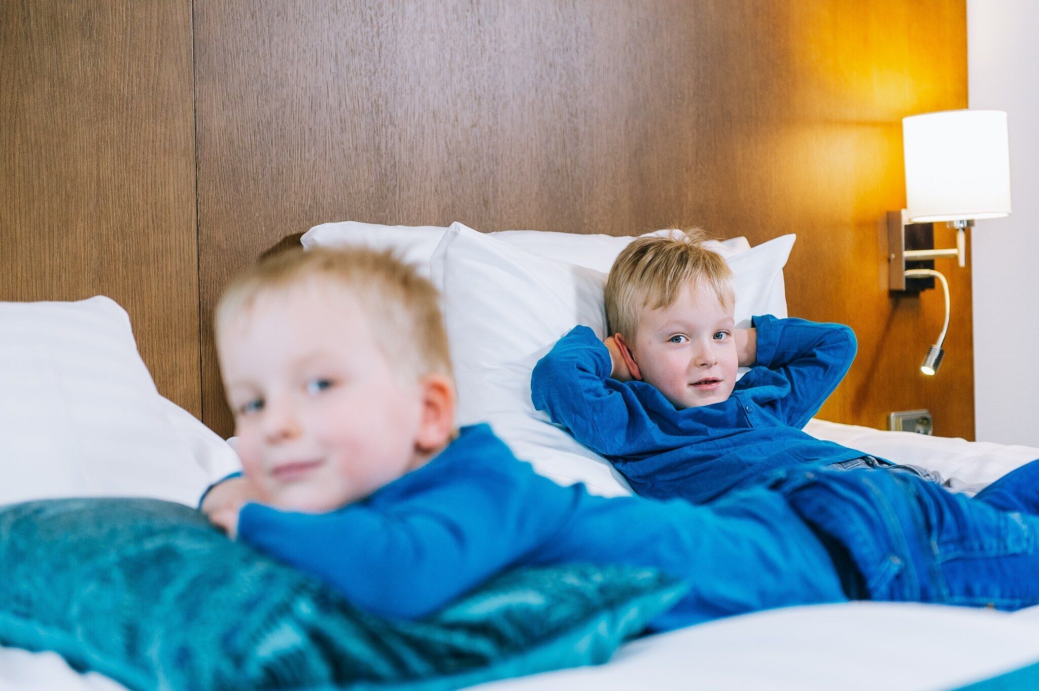 Twee kinderen liggen in bed met witte kussens en een houten hoofdeinde.