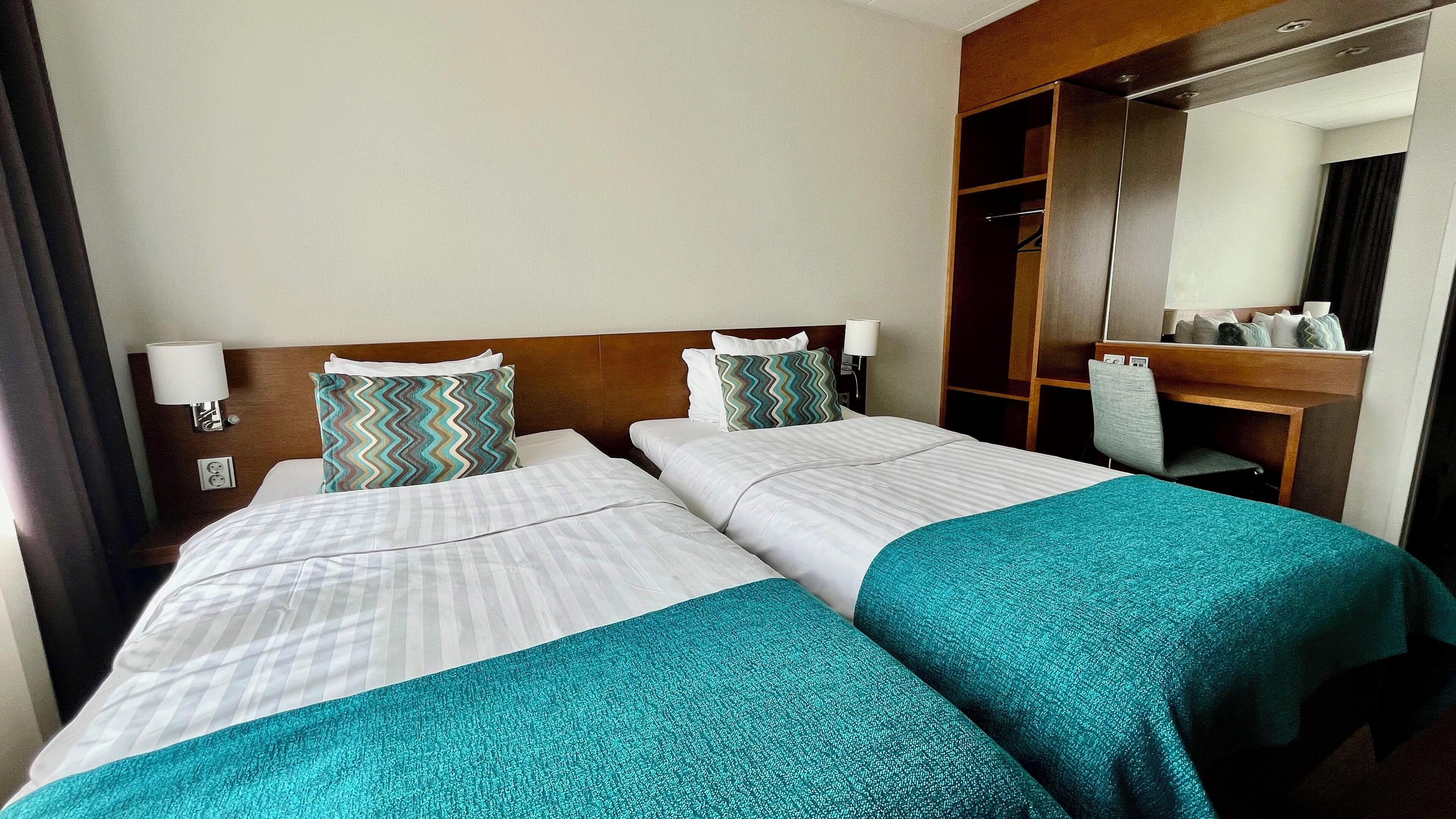Habitación de hotel con dos camas, cabecera de madera, un espejo y un escritorio con silla.