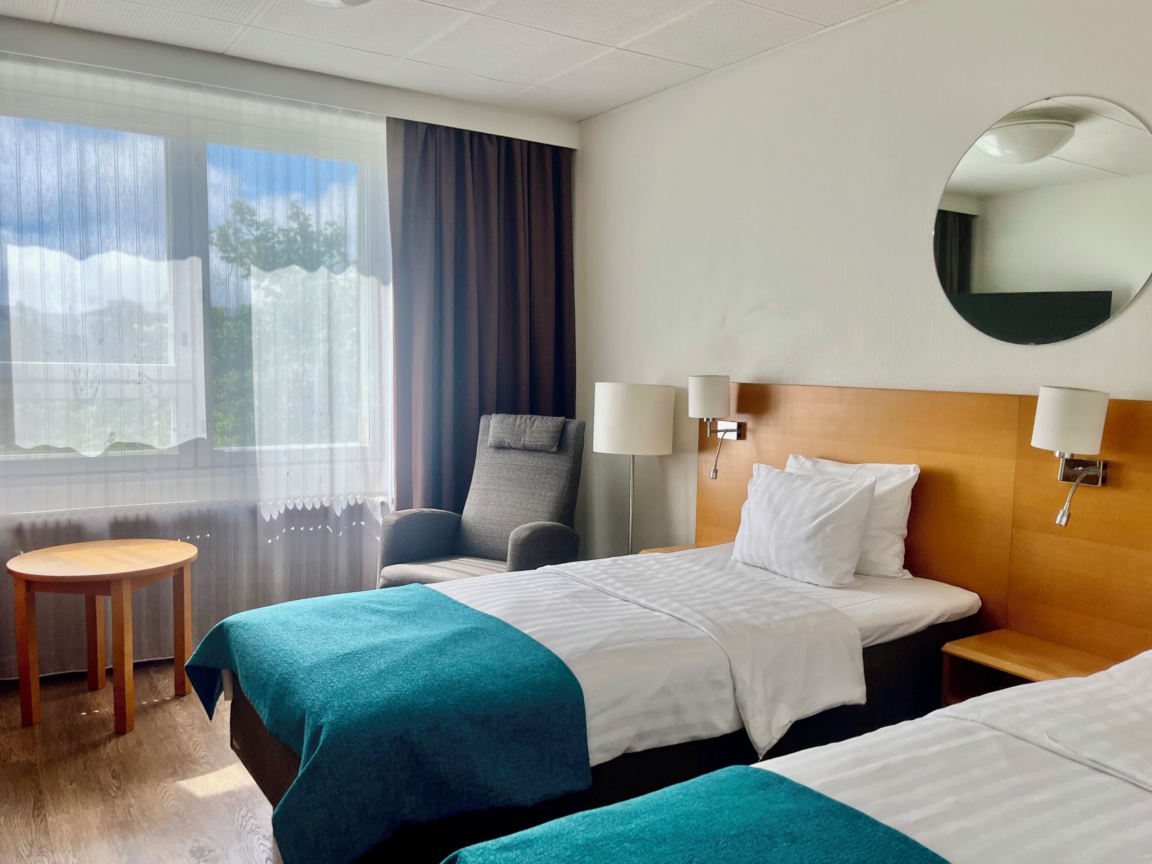 A hotel room with two beds, a chair, a small table, and a large window with curtains.