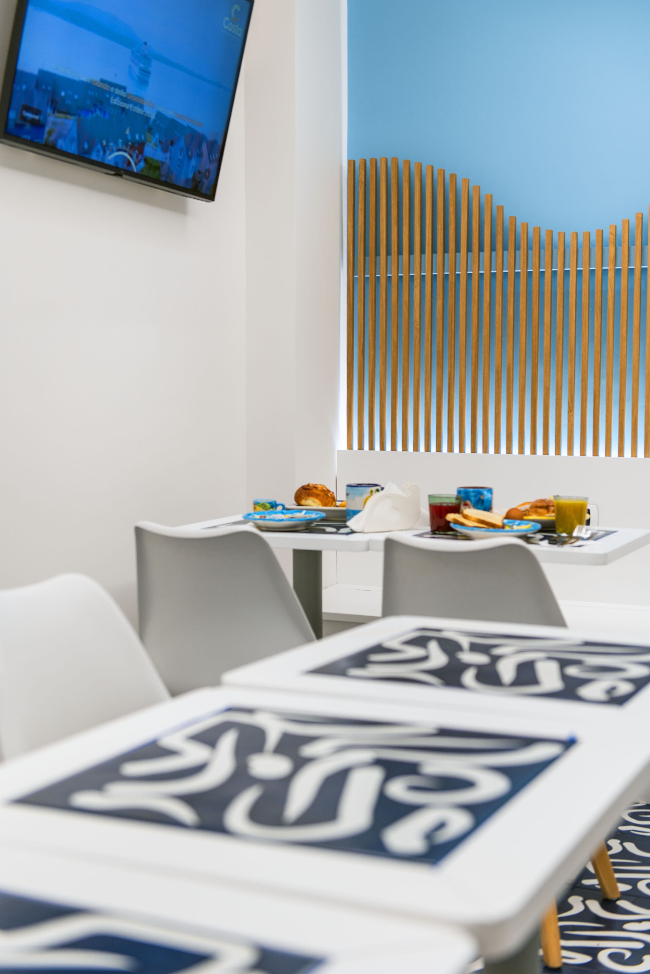 A modern dining area with a TV mounted on the wall, a wooden slat partition, and a table set with plates, glasses, and a croissant.