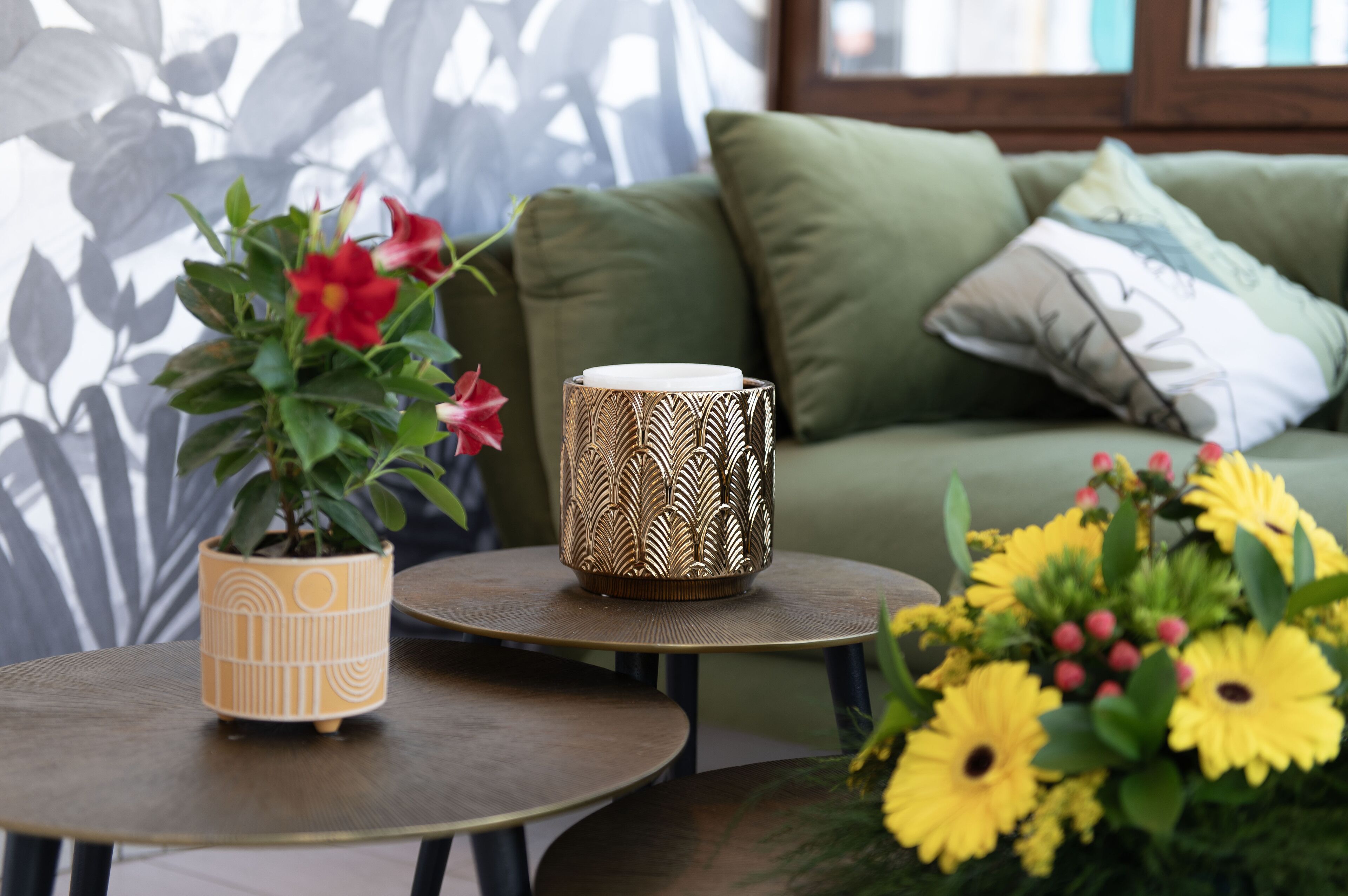 A living room with a green sofa, a small round table with a decorative lamp, and a potted plant with red flowers.