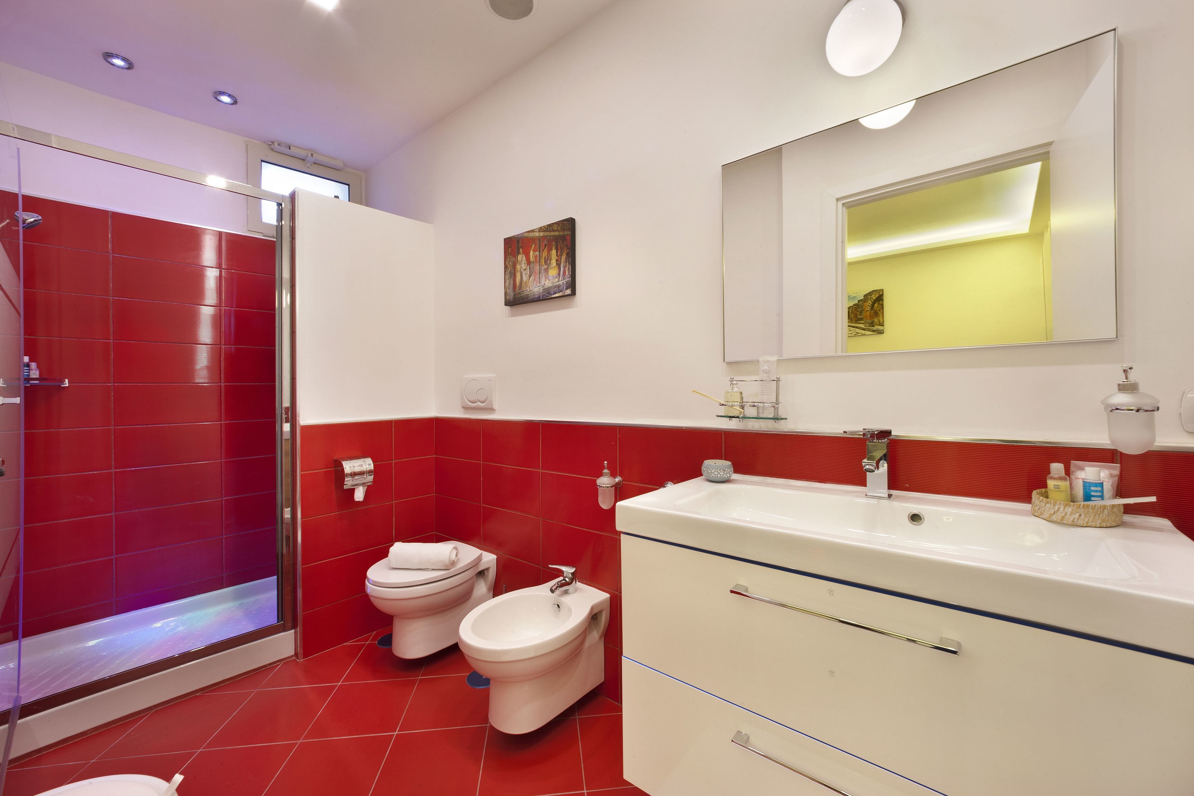 Een badkamer met een rode en witte kleurstelling, met een douche, twee toiletten, een toiletbronschaal, een grote spiegel en een wastafelmeubel met een wastafel.