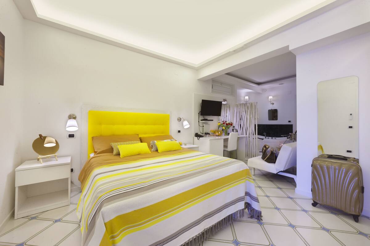 A modern hotel room with a large bed, a desk, a chair, a suitcase, and a TV.