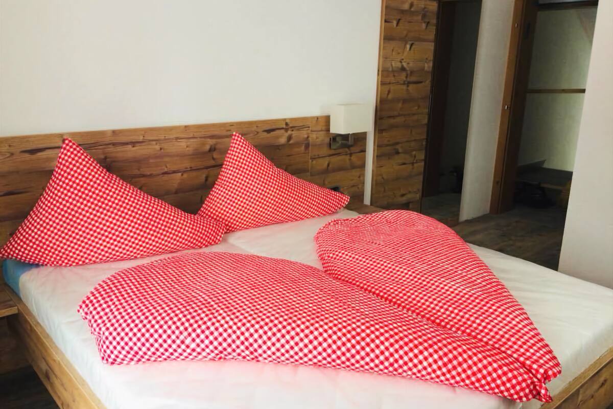 Una cama con colchón blanco y almohadas rojas a cuadros, cabecero de madera y una mesita de noche con lámpara.