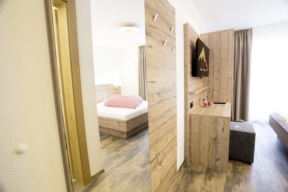 Una habitación de hotel moderna con piso de madera, una cama con almohada a cuadros rojos y blancos, un pequeño escritorio de madera y un televisor montado.