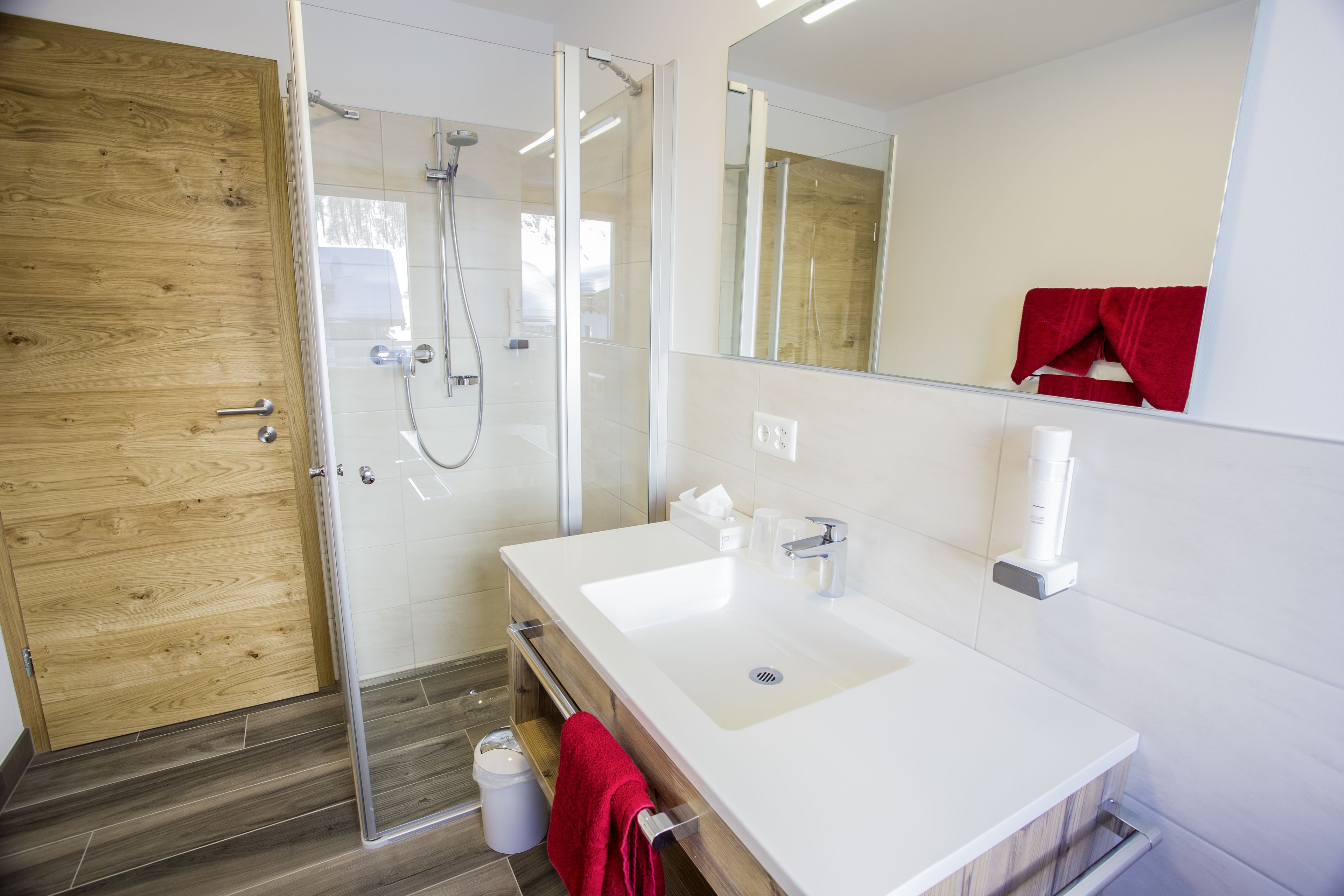 Une salle de bain moderne avec une porte en bois, une douche en verre, un lavabo blanc et une serviette rouge suspendue à un porte-serviettes.