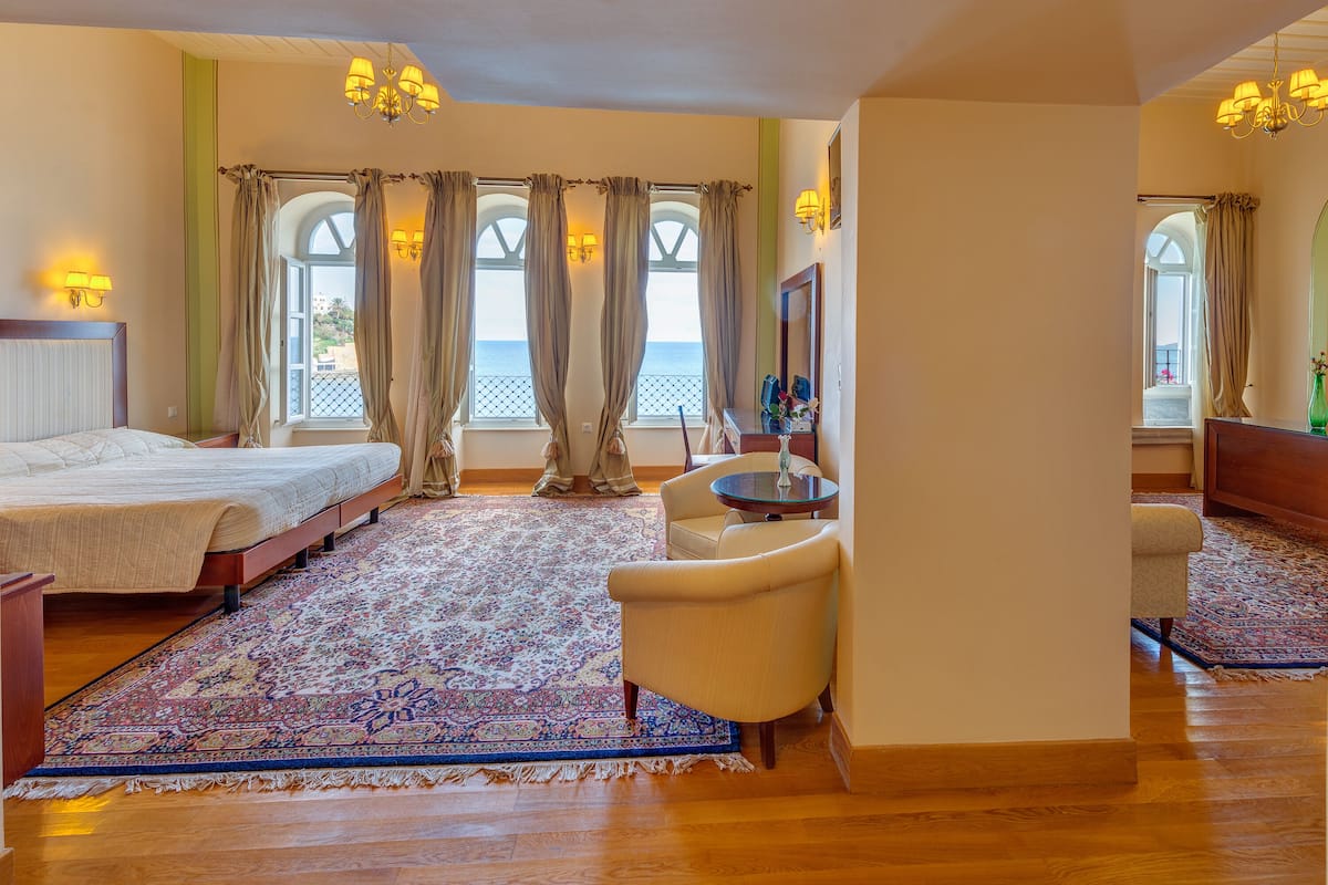 Habitación de hotel con una cama grande, una zona de estar con una mesa y una silla, vista al mar a través de amplios ventanales y un candelabro.