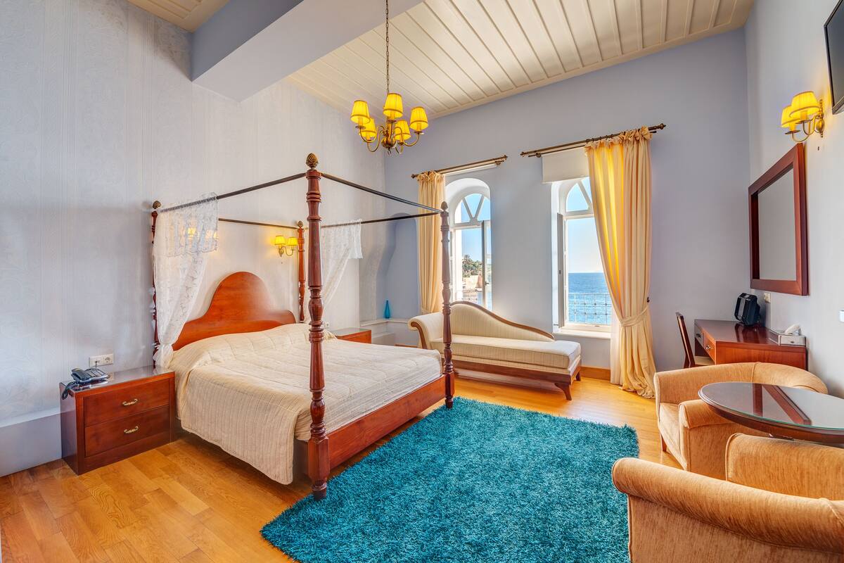A bedroom with a four-poster bed, a sitting area with a sofa and armchair, a small table, and a view of the sea through a window.