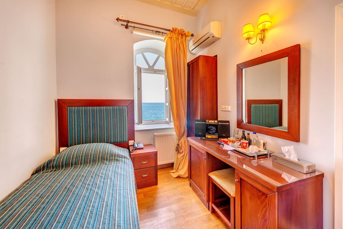 Habitación de hotel pequeña con cama, escritorio de madera, espejo, ventana con cortinas y vista al mar.