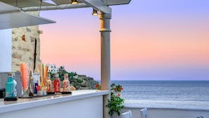 A balcony with a view of the ocean at sunset, a bar area with various bottles, and a small table with a chair. - Apollonion Palace Hotel (Syros)