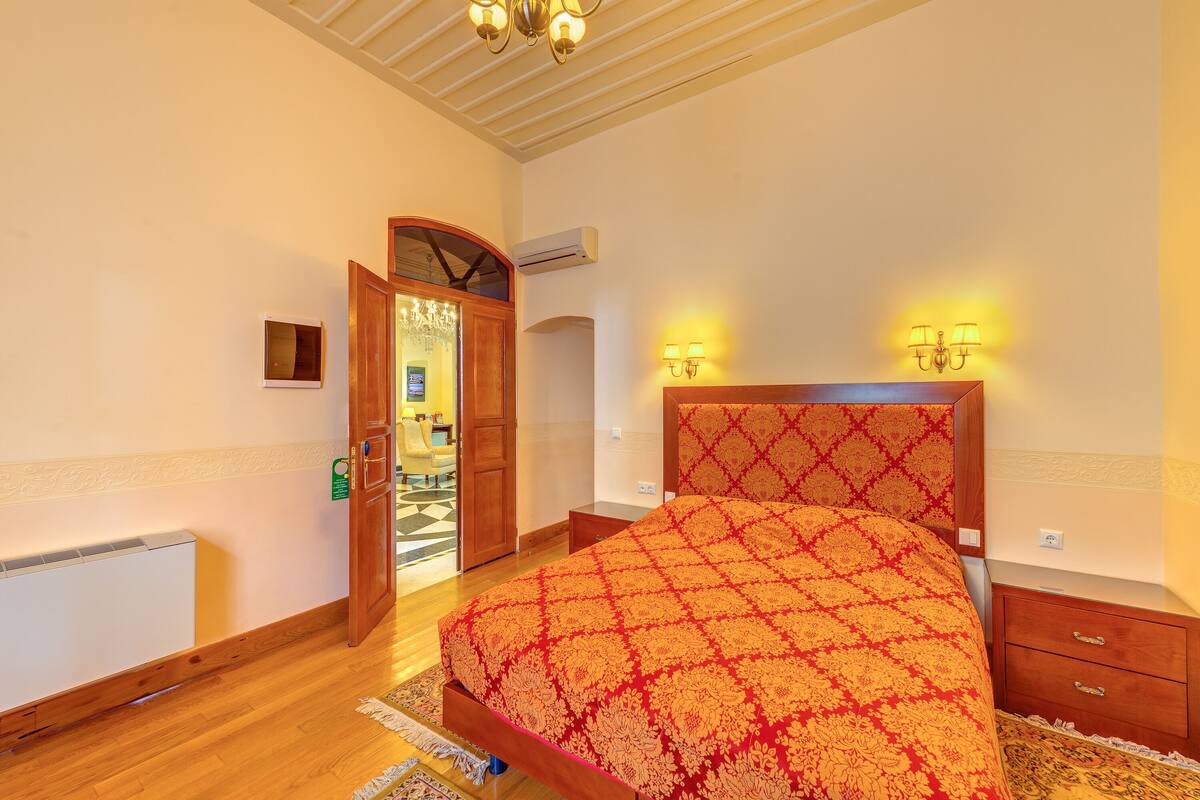 A bedroom with a large bed, wooden headboard, bedside lamps, a radiator, and a door leading to another room.