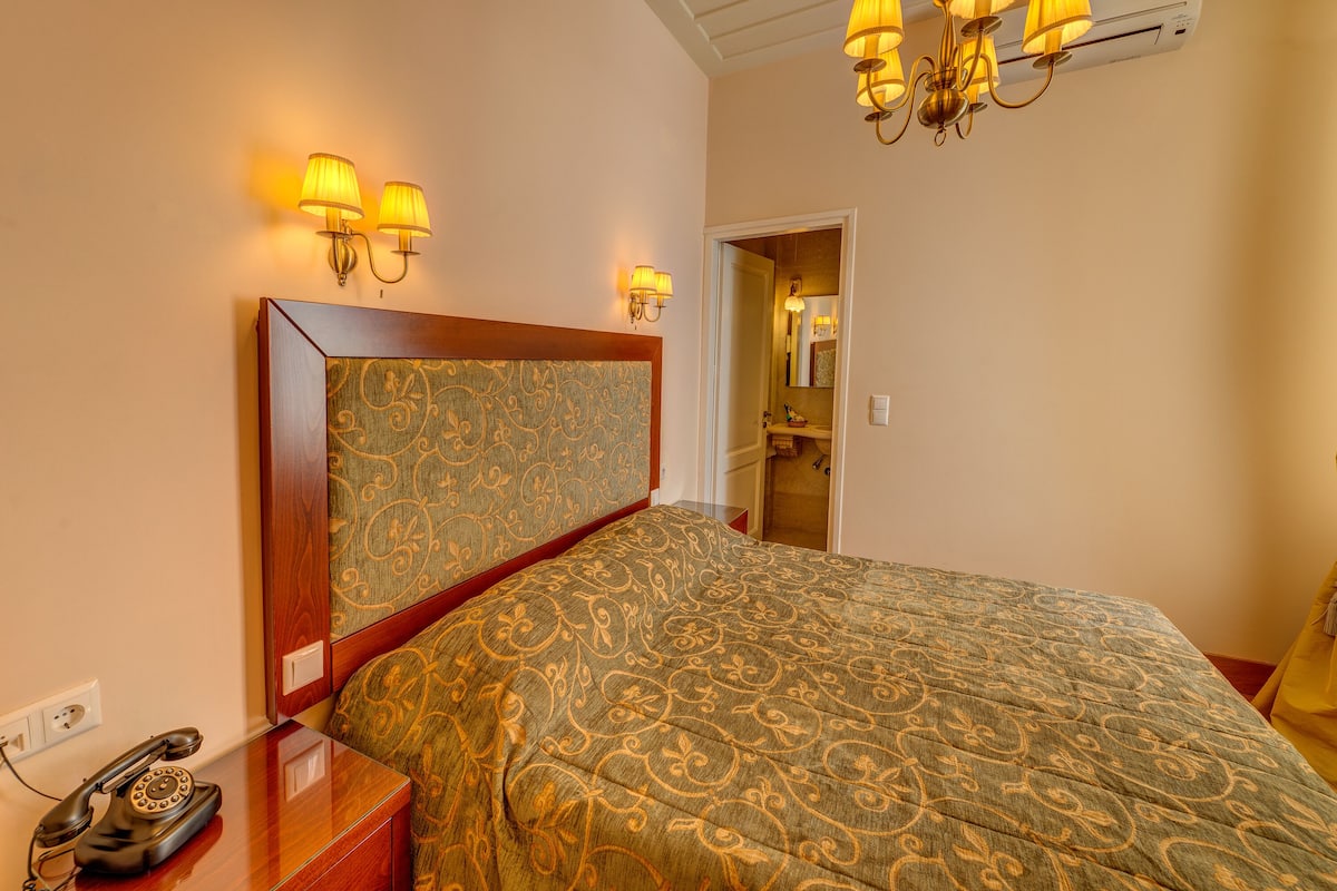 A hotel room with a patterned bedspread, a wooden bedside table with a rotary phone, wall-mounted lamps, and a chandelier.