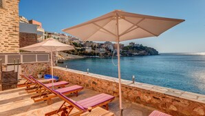 A seaside terrace with lounge chairs and umbrellas, a stone wall, and a view of the sea and buildings. - Apollonion Palace Hotel (Syros)