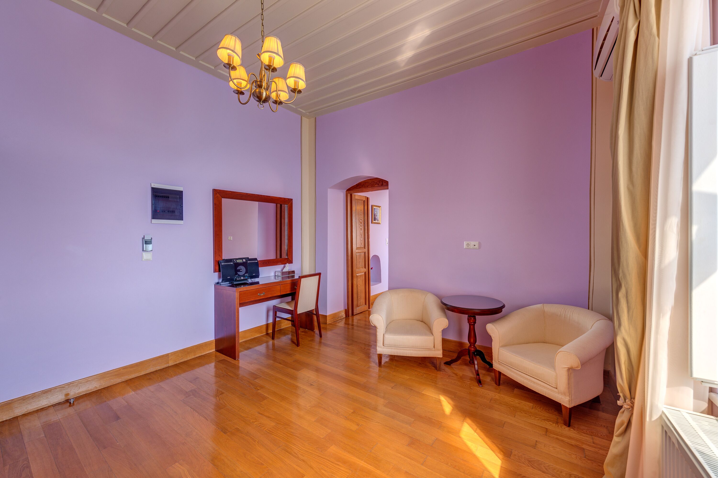 A room with wooden flooring, a desk with a chair, a sofa, and a chandelier.