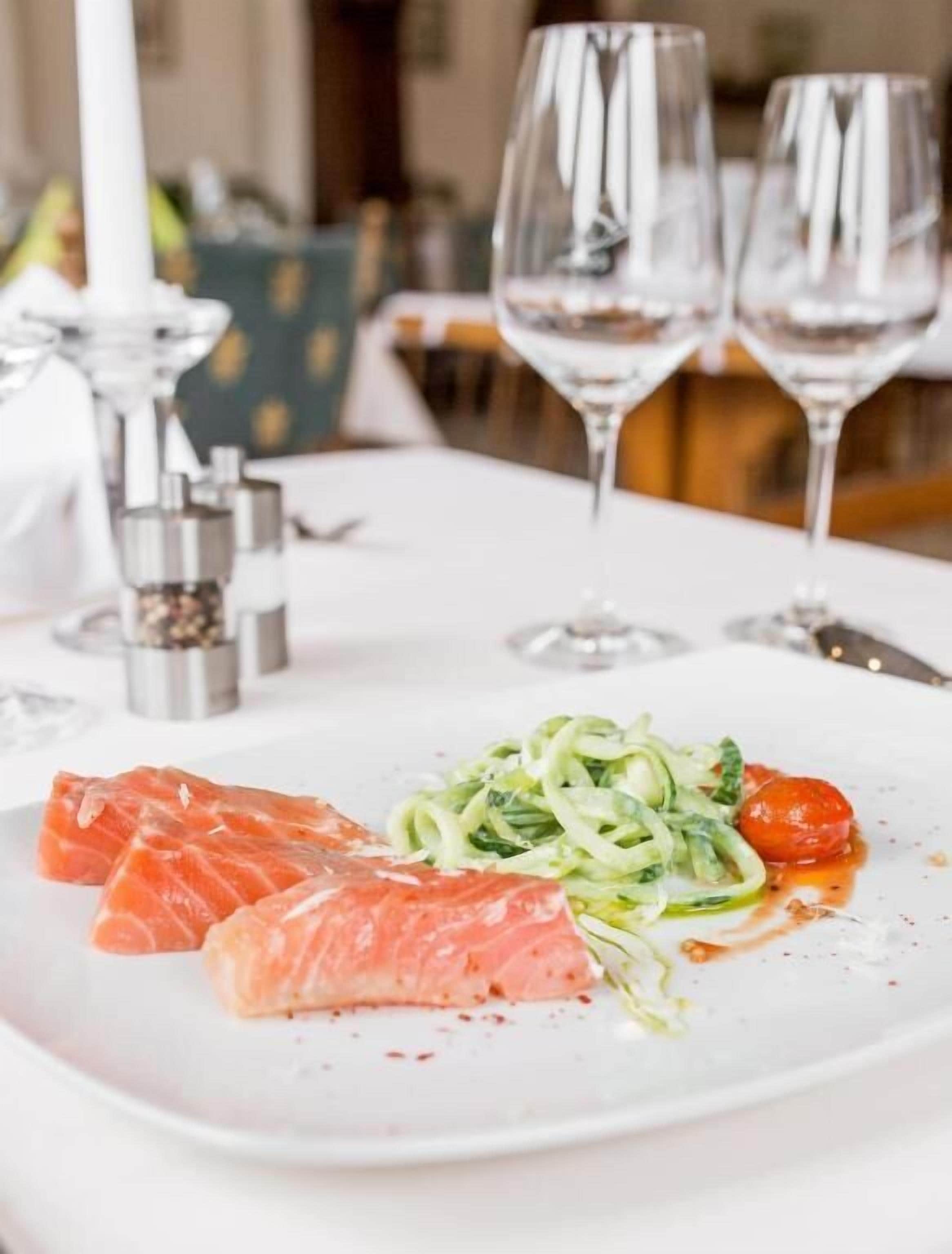 Une assiette blanche contenant du saumon, des nouilles de courgettes et des tomates cerises, posée sur une table de restaurant.