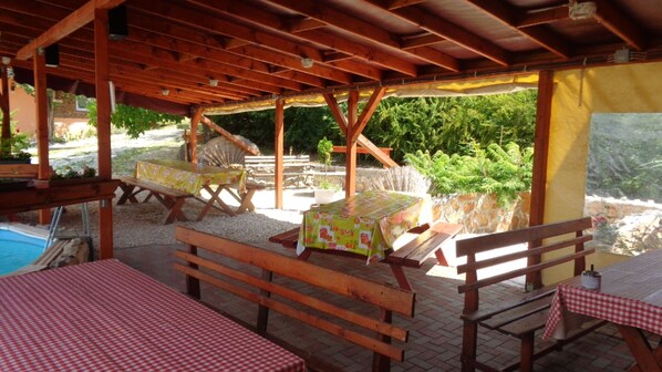 An outdoor patio area with wooden tables and benches, covered by a wooden roof structure.
