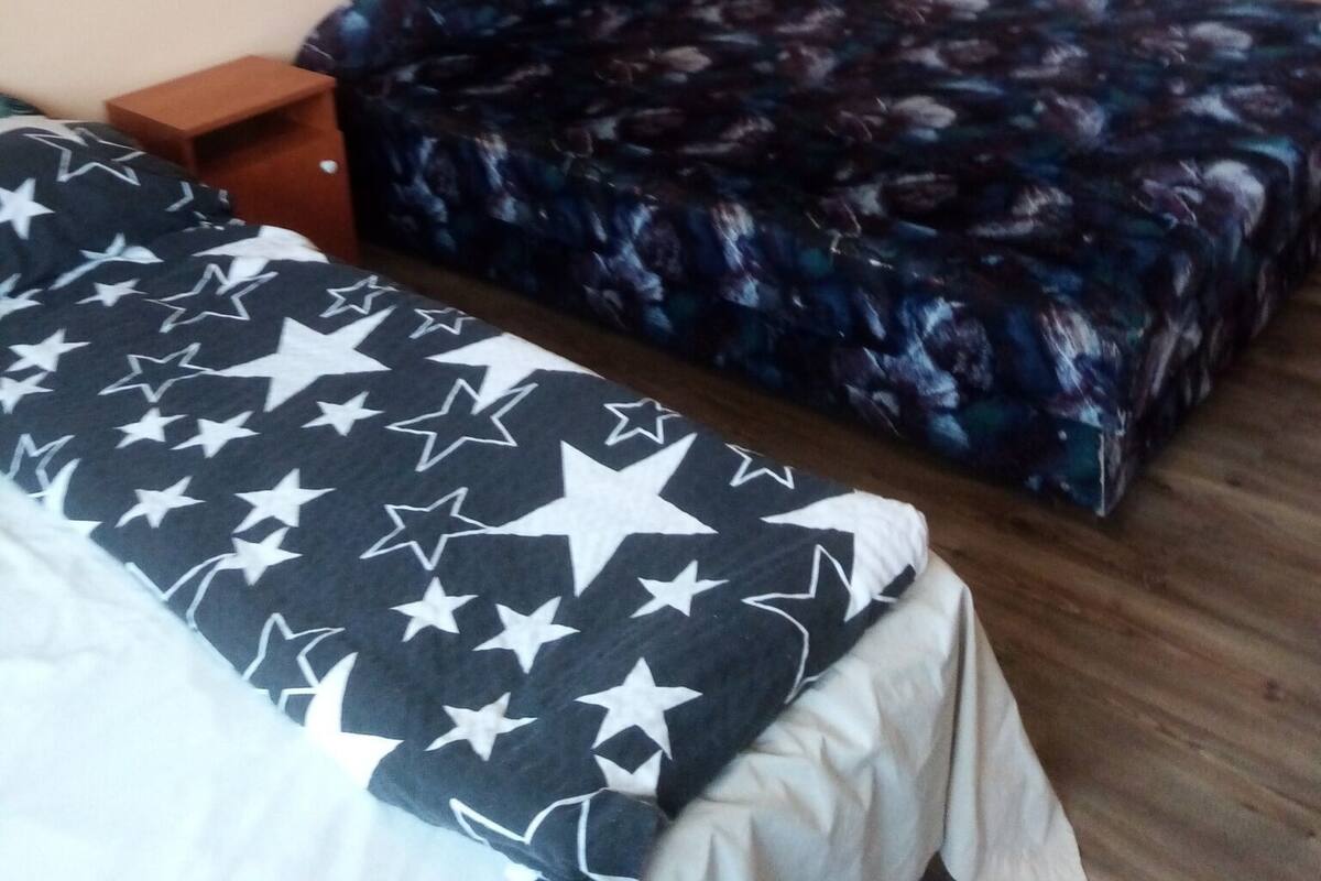A room with two beds, one with a star-patterned bedspread and the other with a floral-patterned one. There are two wooden nightstands, one on each bed.