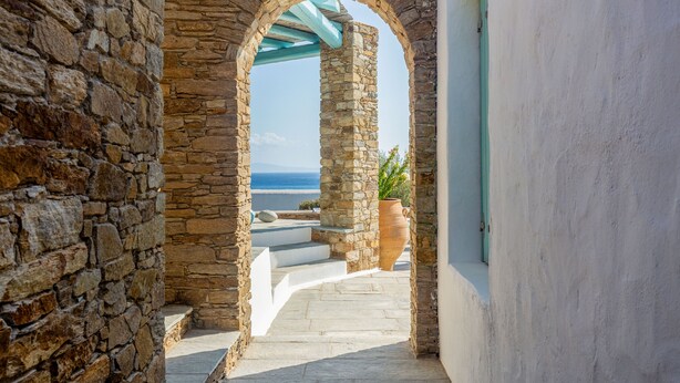 A stone corridor with a view of the sea, a tiled floor, and a large terracotta pot. - Magganari View Villa - Discover Ultimate Summer Happiness! (Magganari Ios Island)